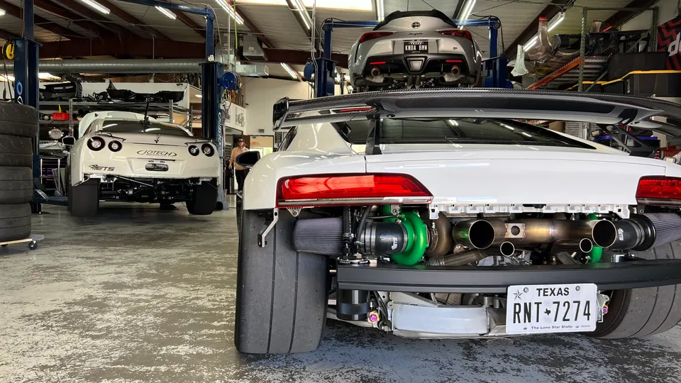 A white Audi R8 with exposed twin turbochargers and custom exhaust in a performance tuning shop, with a Nissan GT-R and Toyota Supra in the background.