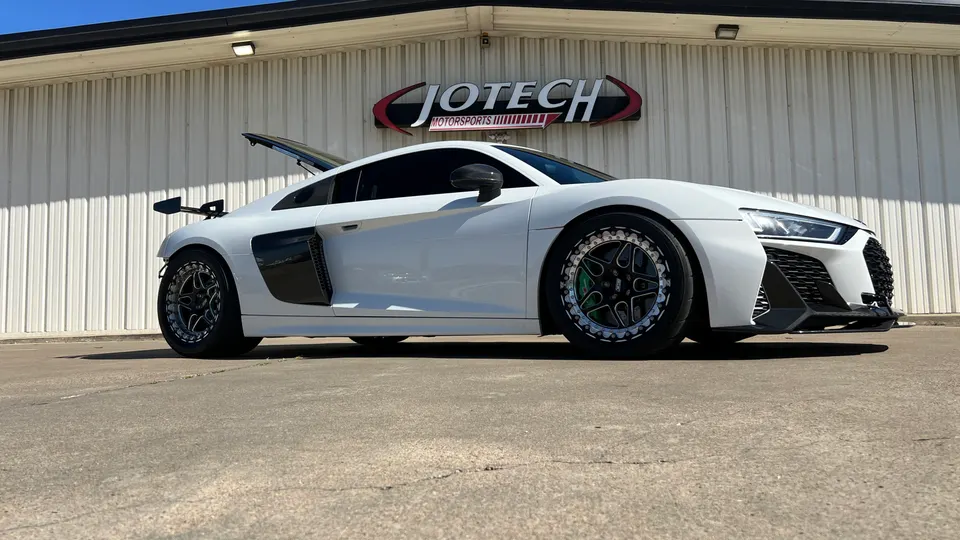 A white Audi R8 performance car with aftermarket wheels and a rear wing parked outside Jotech Motorsports.