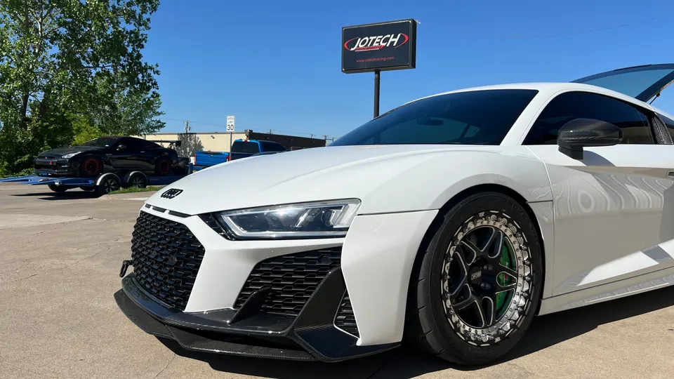 A white Audi R8 with aftermarket wheels and a carbon fiber front splitter parked outside Jotech, with a black Nissan GT-R on a trailer.