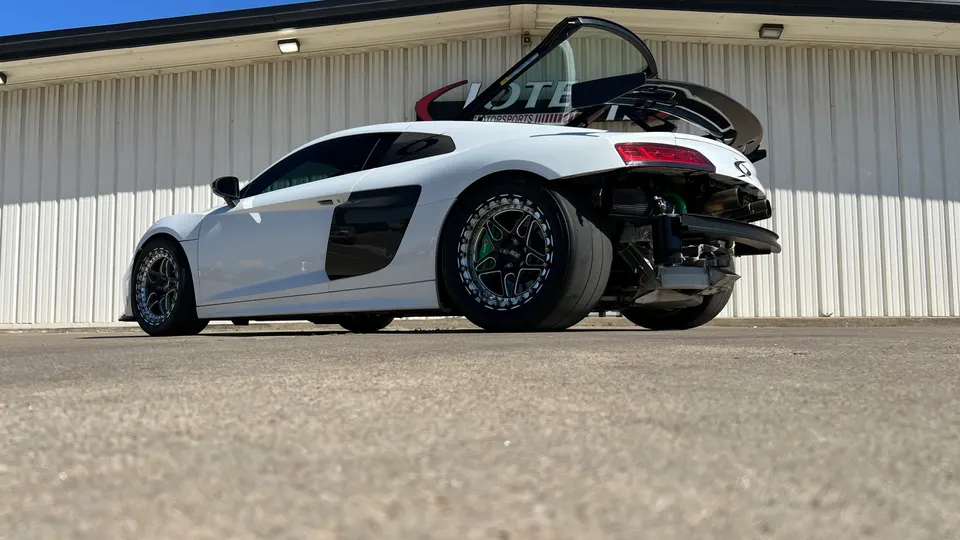 White Audi R8 drag car with aftermarket wheels and exposed exhaust system parked outside a building.