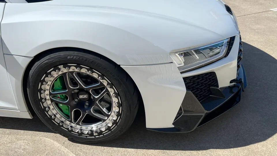 A close-up of the front wheel and fender of a white Audi R8, showcasing custom black wheels with chrome hardware, green brake calipers, and a carbon fiber front