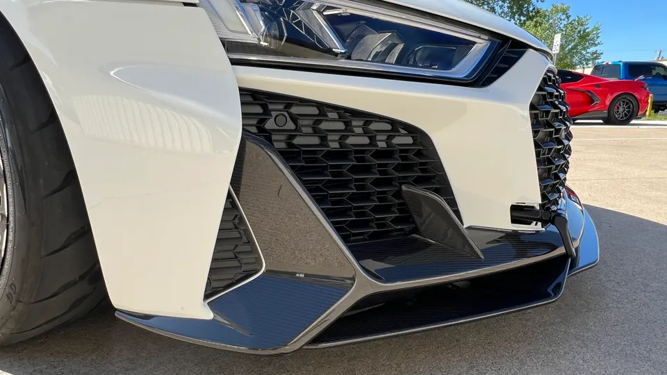 Close-up of a white Audi RS6 front bumper with carbon fiber splitter and honeycomb grille.