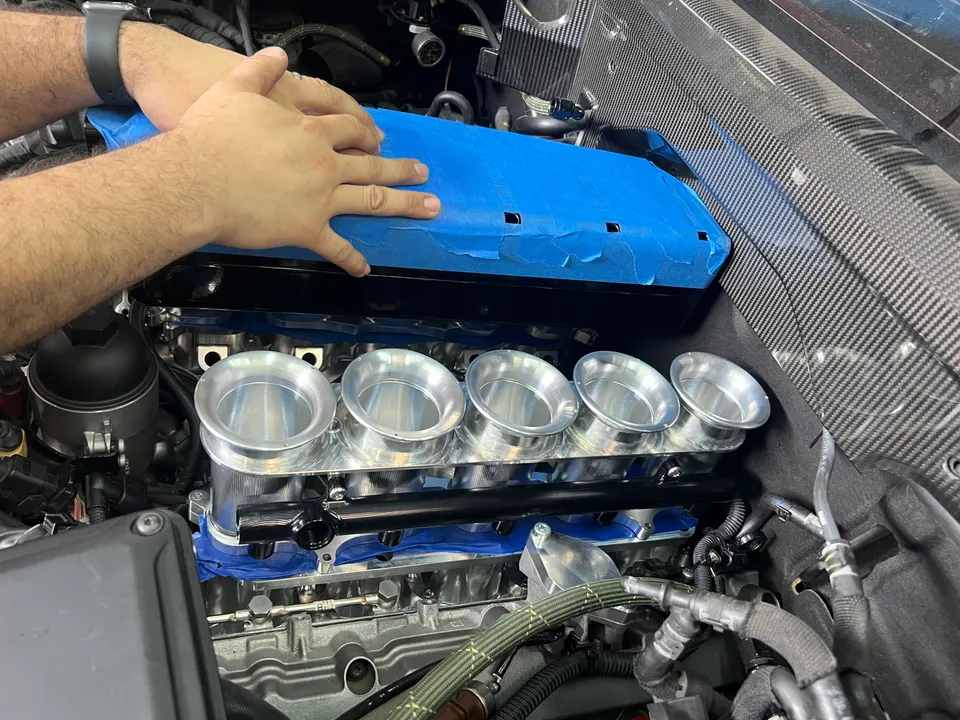 A person's hands working on an engine bay, installing an aftermarket intake manifold with velocity stacks.