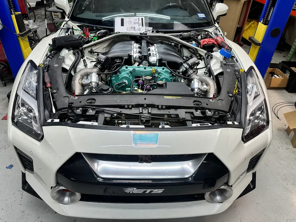 White Nissan GT-R with its hood open in a shop, revealing a heavily modified twin-turbo engine bay.