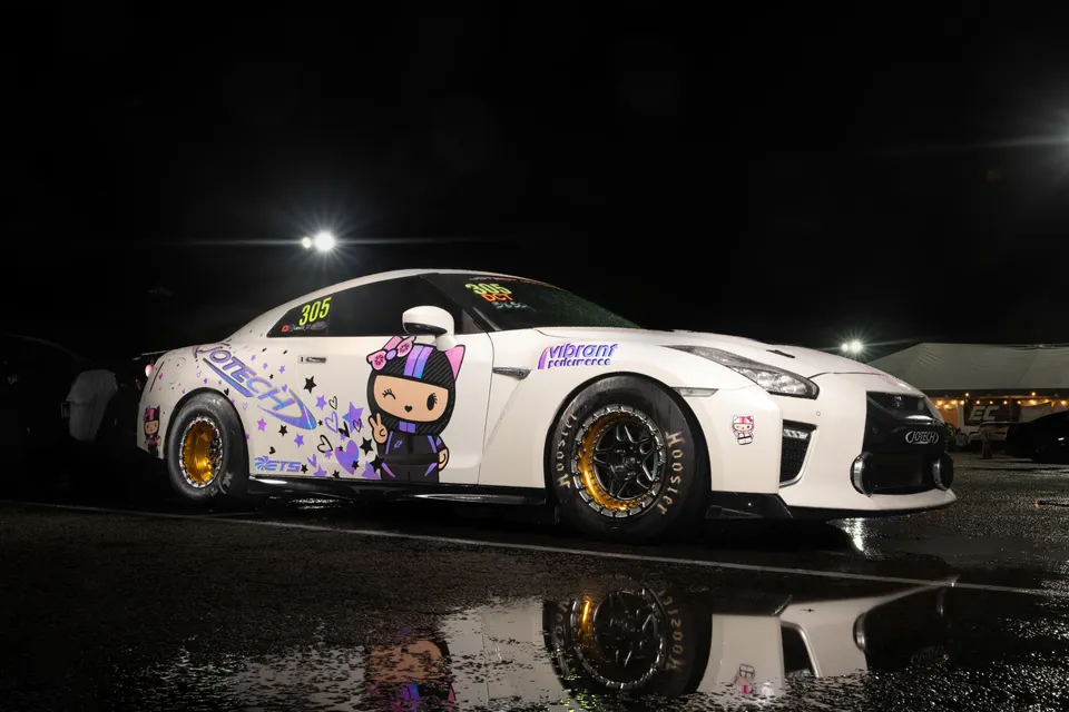 A white Nissan GT-R drag car with custom purple livery and gold drag wheels on a wet track at night, ready for a race.