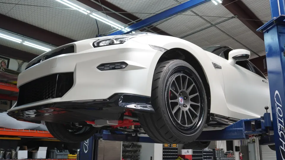 A white Nissan GT-R on a lift in a Jotech Motorsports shop, showcasing its underside, wheels, and suspension.