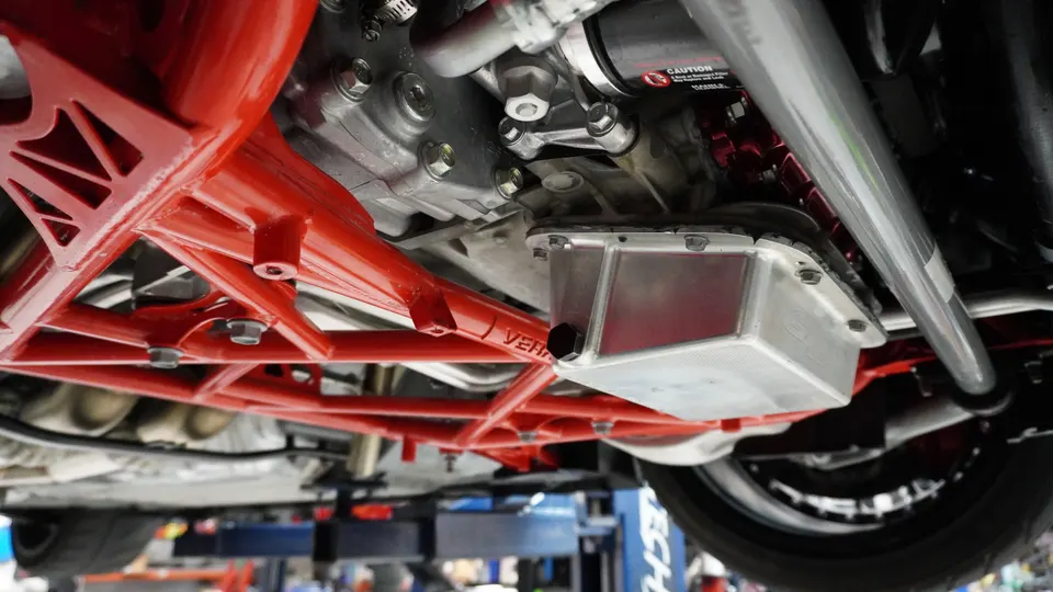Underside view of a performance car showing a red aftermarket subframe, custom oil pan, and engine components on a lift.