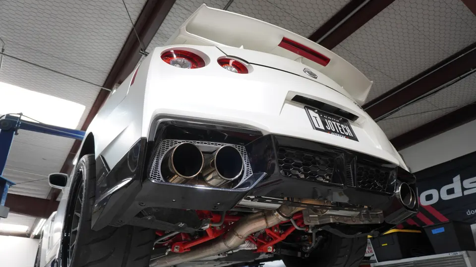 A white Nissan GT-R on a lift in a shop, showcasing its aftermarket exhaust system and underbody.