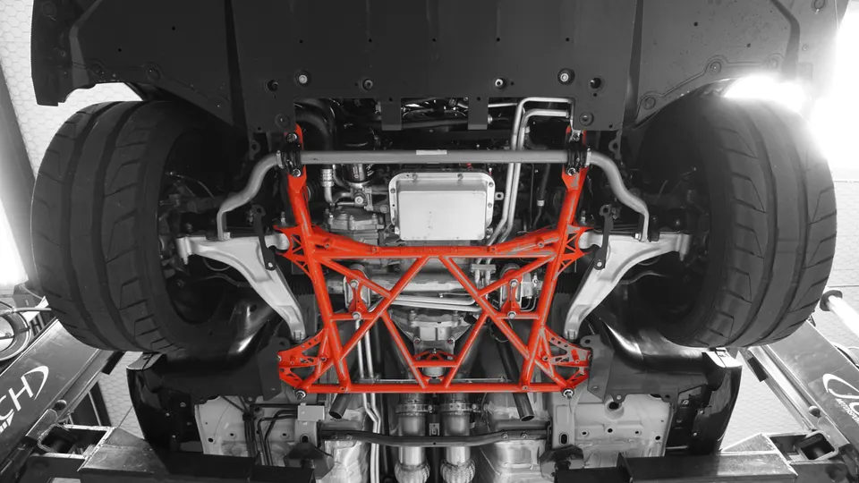 Underside view of a performance car on a lift, showcasing a vibrant red aftermarket chassis brace, suspension, and wide tires.