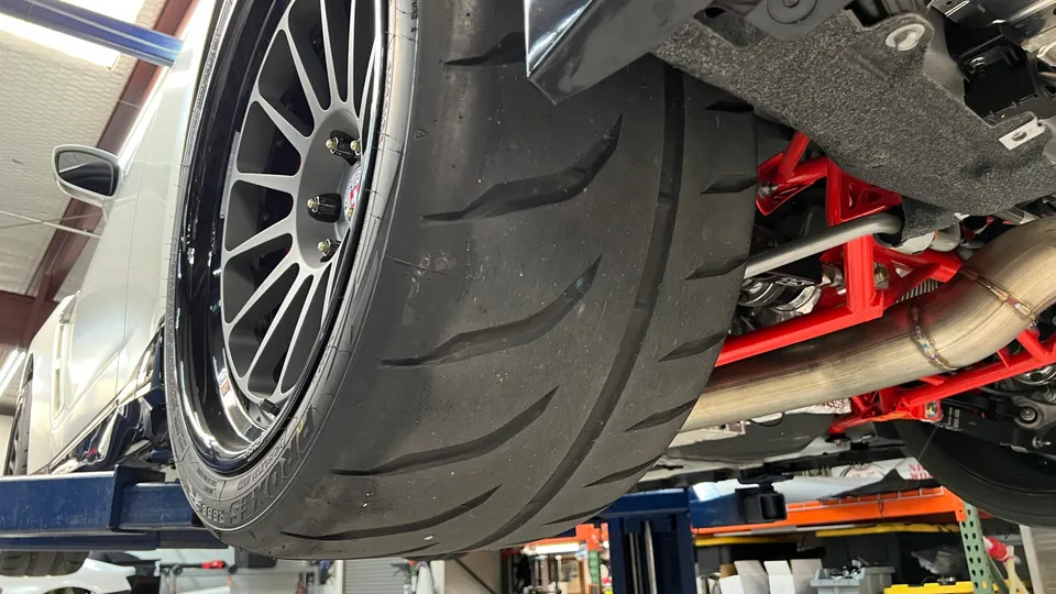 Car on a lift showcasing a racing slick tire, aftermarket wheel, red suspension, and custom exhaust in a shop.