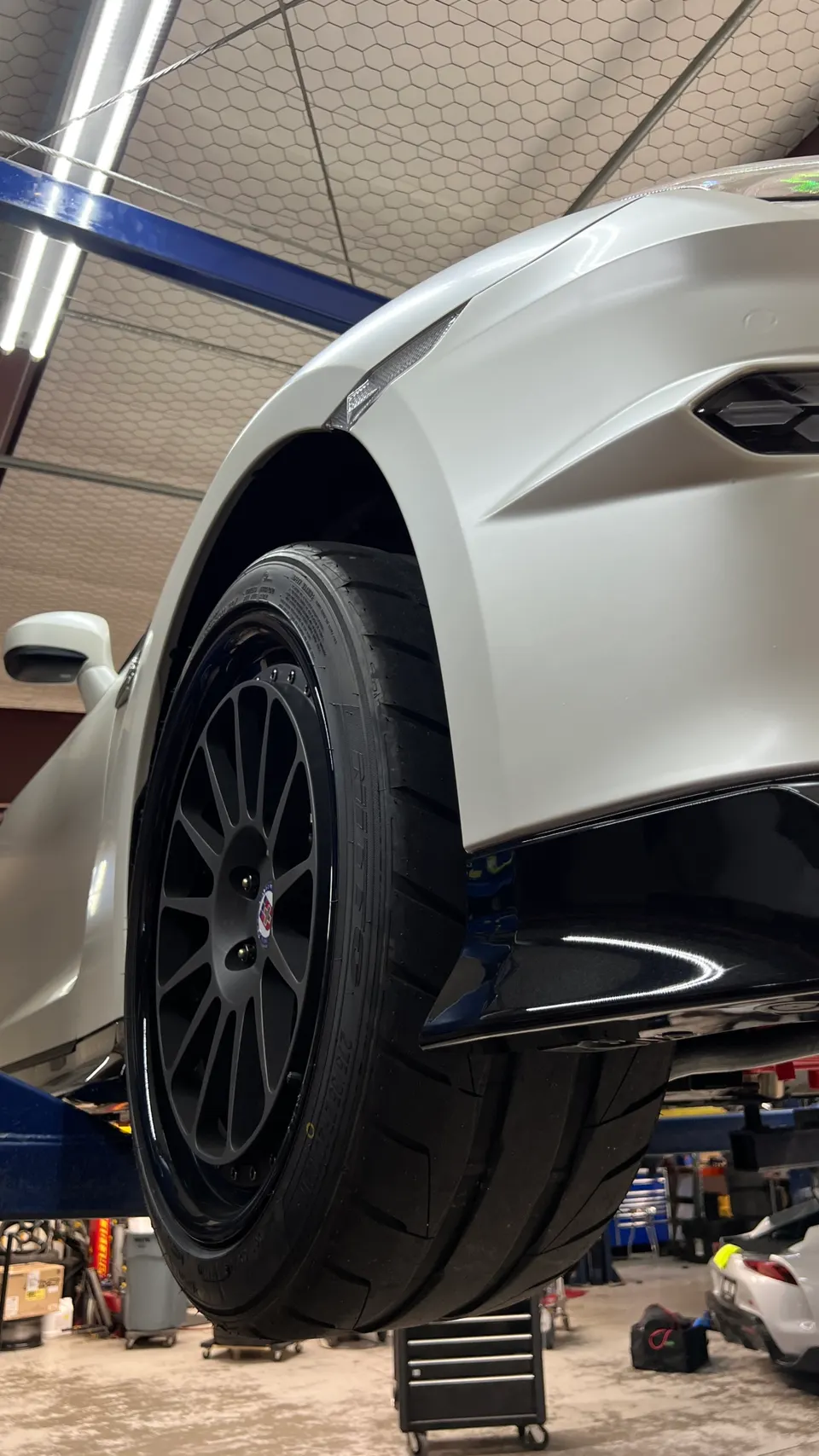 A white Nissan GT-R on a lift in a shop, showcasing its aftermarket wheel, performance tire, and suspension.