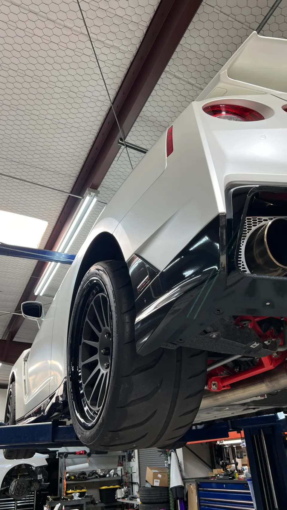 A white Nissan GT-R on a lift in a performance shop, showcasing aftermarket wheels, performance tires, and custom suspension.