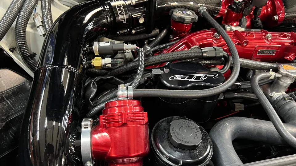 A close-up of a Nissan GT-R engine bay featuring a red blow-off valve, black charge pipe, and red valve covers.