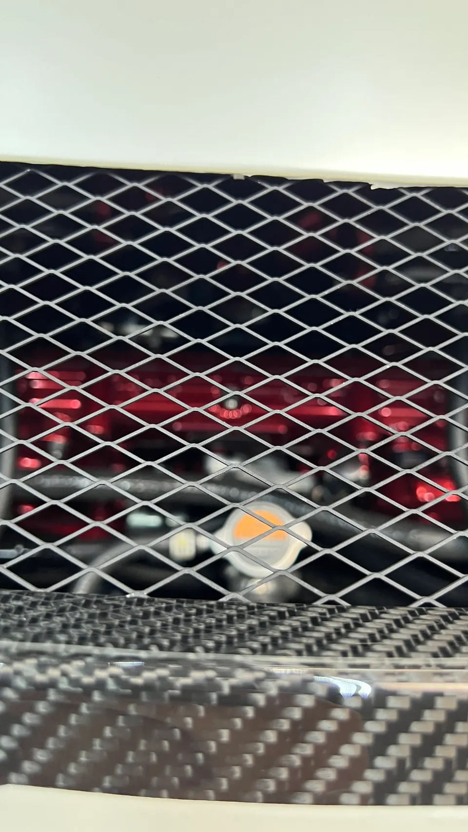 Close-up view of a car's engine bay through a grille, showing a red component and carbon fiber trim.