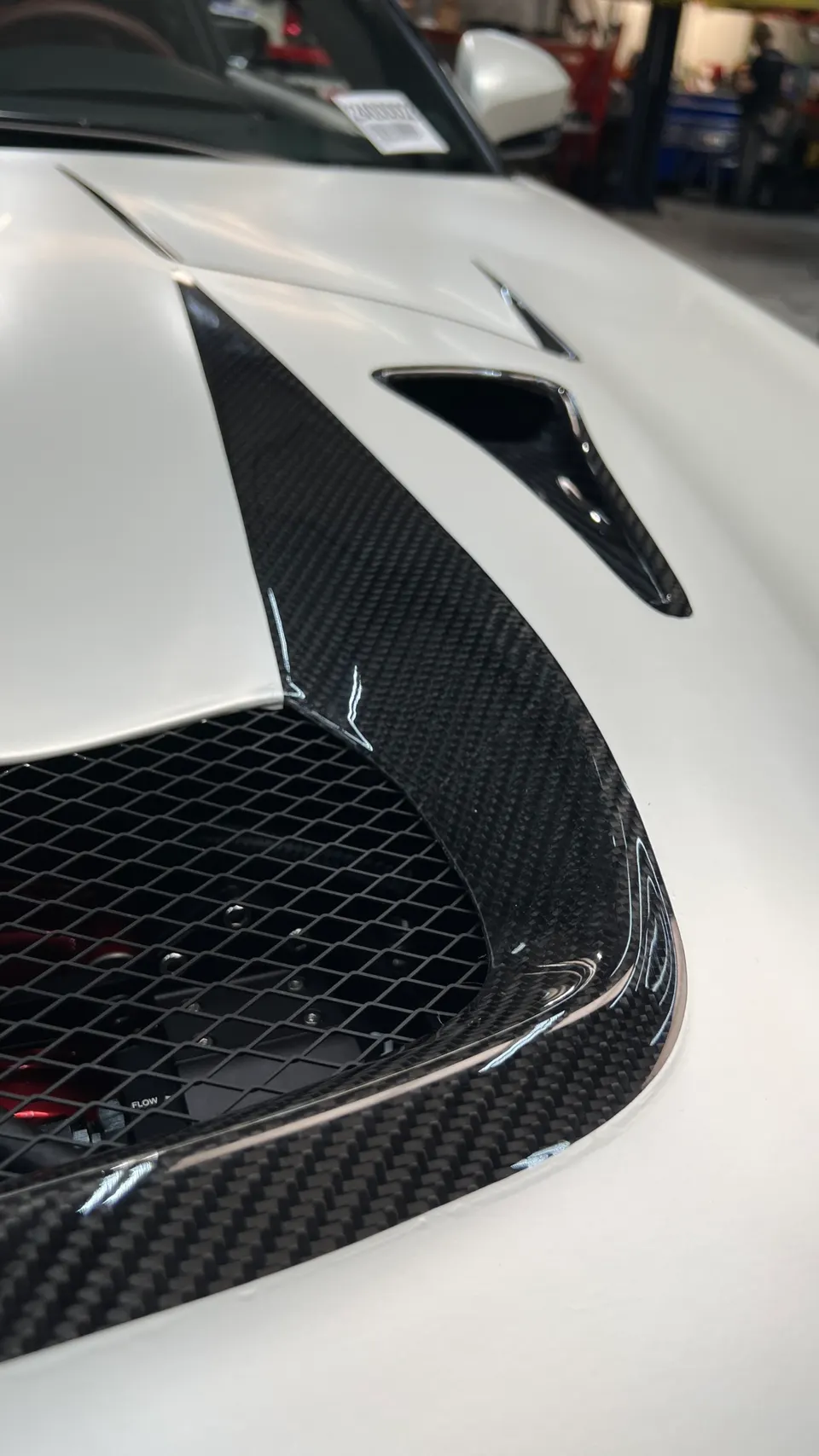 Close-up of a white car's hood with carbon fiber vents and a mesh grille in a shop.