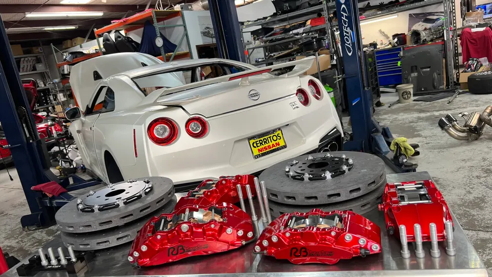 White Nissan GT-R on a lift in a shop, with large red performance brake calipers and carbon ceramic rotors on a table.