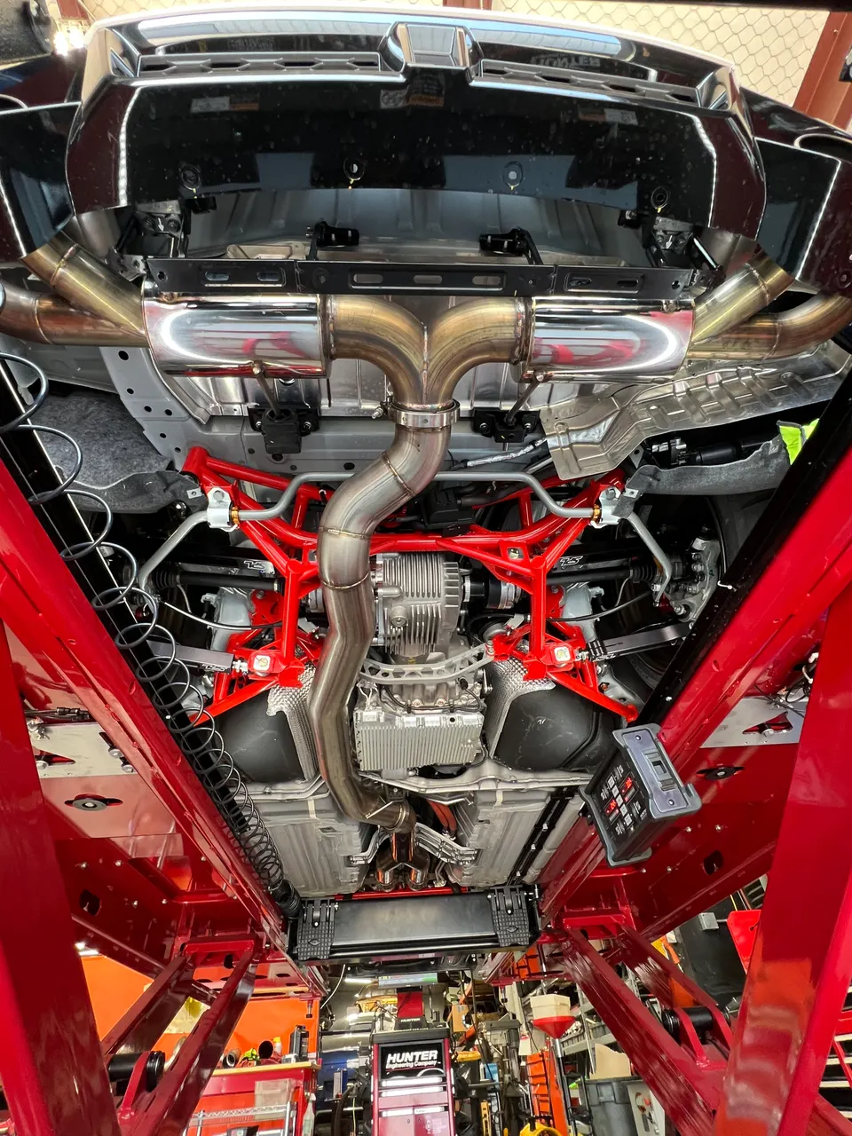 Underside view of a performance car on a red lift, showcasing a custom exhaust system, red suspension components, and differential.
