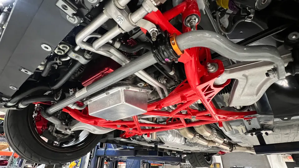 A detailed view of a car's underside on a lift, showcasing red aftermarket suspension components, oil pan, and exhaust system.