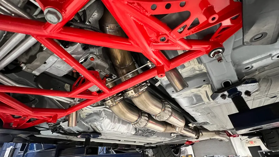 Detailed view of a car's underside showing a custom exhaust system with flex pipes and a red chassis brace in a shop.