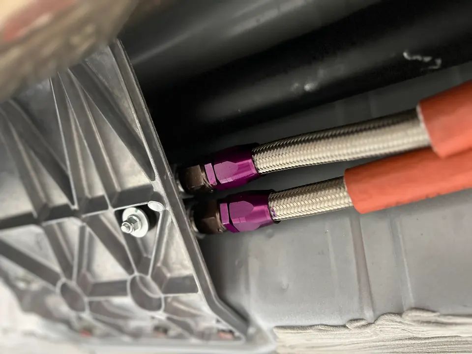 Close-up of braided performance fuel lines with purple AN fittings connected to a car's oil or transmission pan.