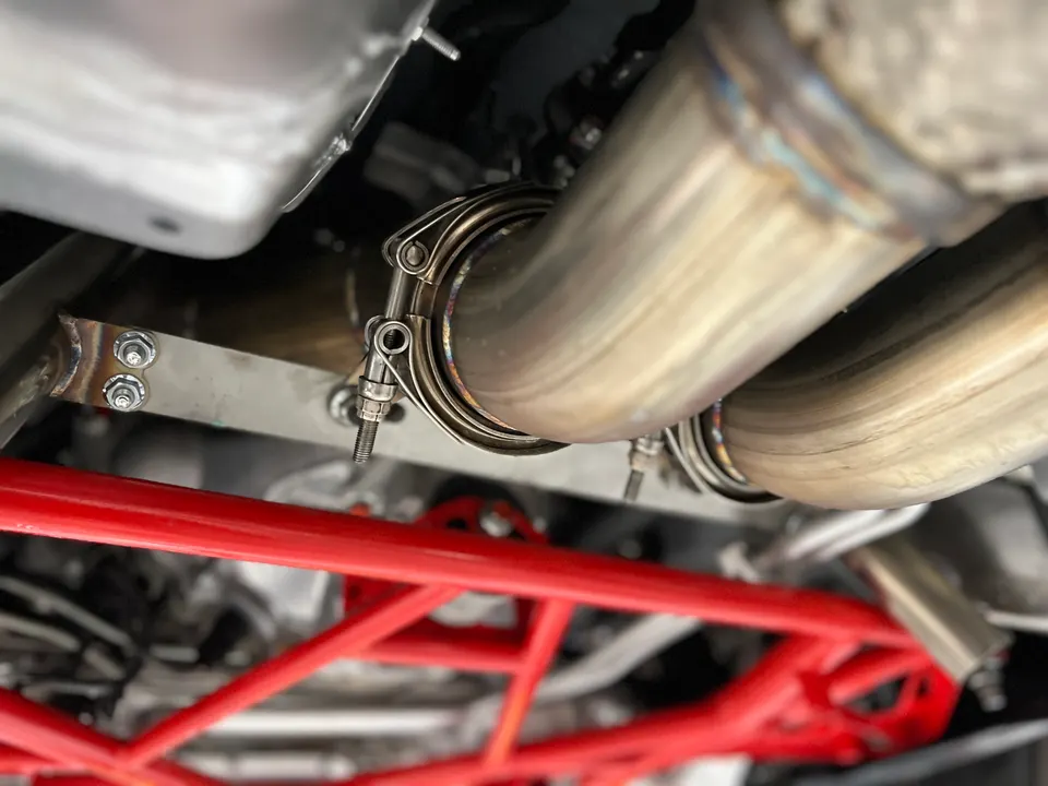 Close-up view of a custom exhaust system with V-band clamps and a red chassis brace on the underside of a performance car.
