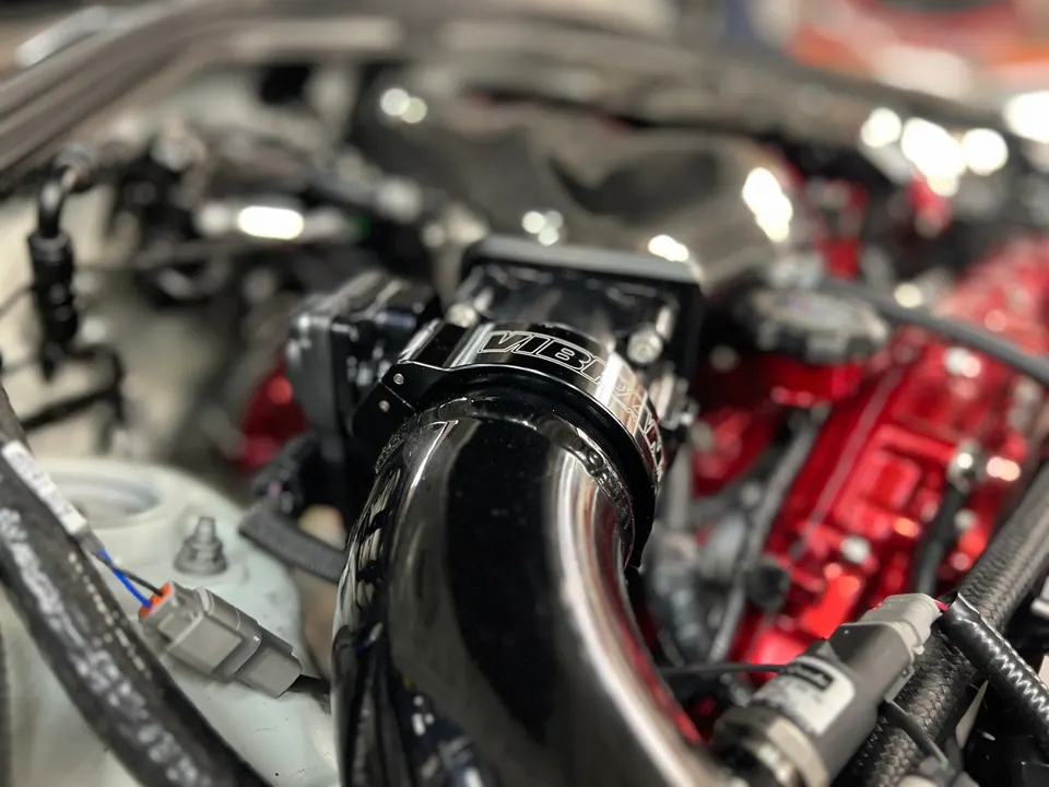 Close-up of a black performance intake pipe with a Vibrant clamp in a car's engine bay, featuring a red engine cover.
