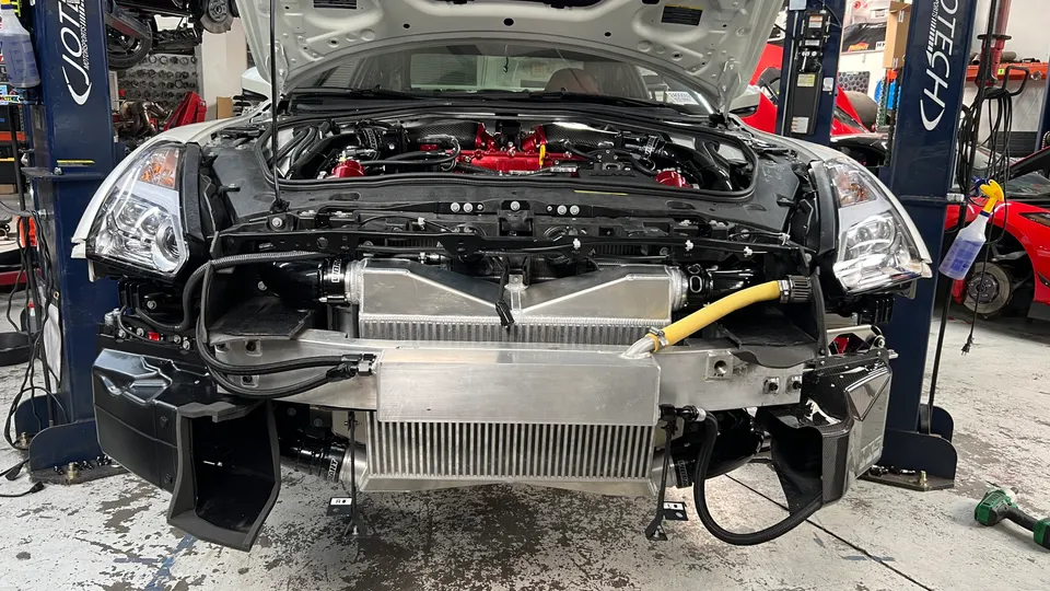 A white Nissan GT-R R35 with its front bumper removed, revealing large aftermarket intercoolers and a red intake manifold in a Jotech Motorsports shop.