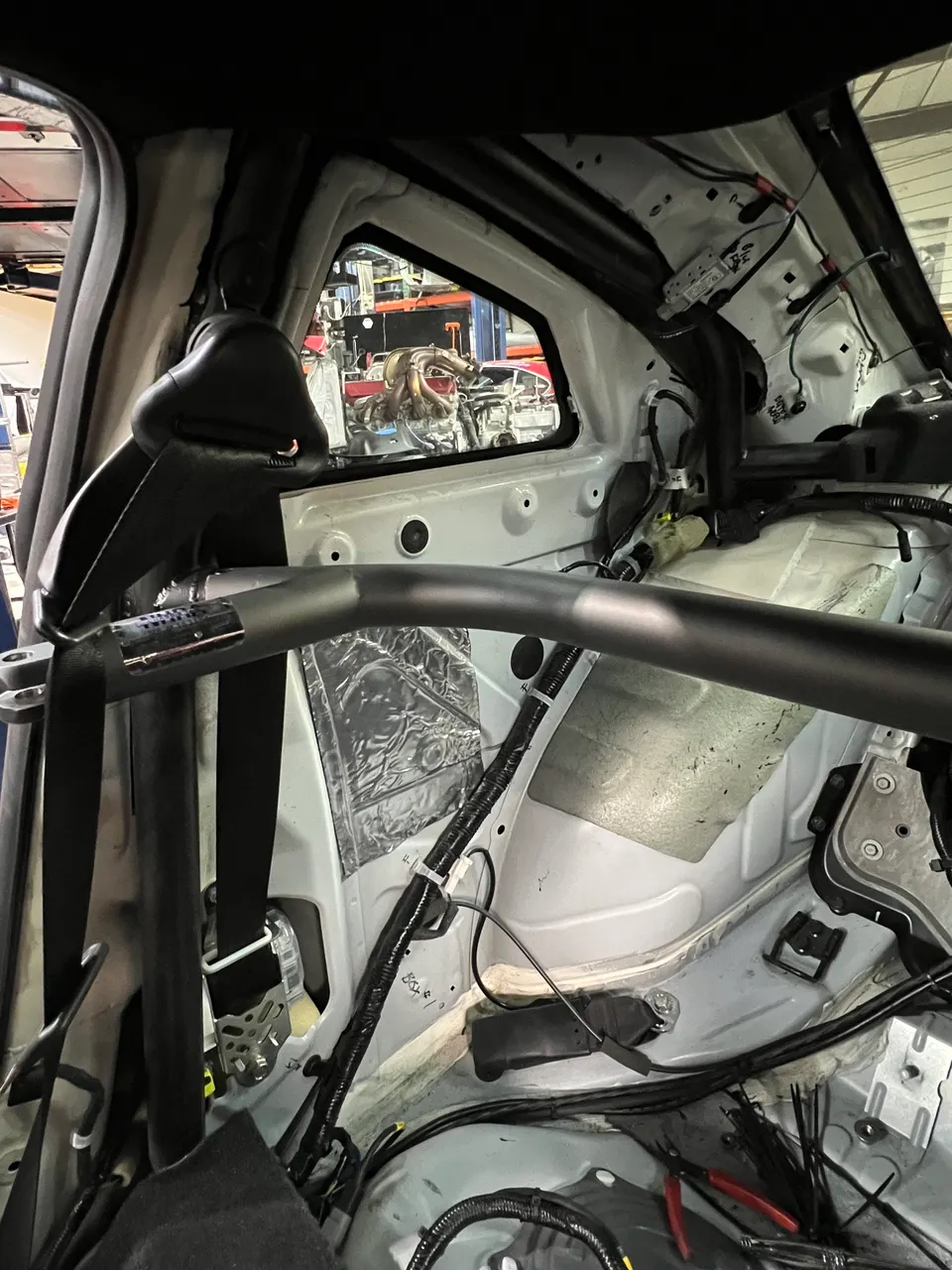 Rear interior view of a performance car with a roll bar installed, exposed wiring, and sound deadening in a tuning shop.