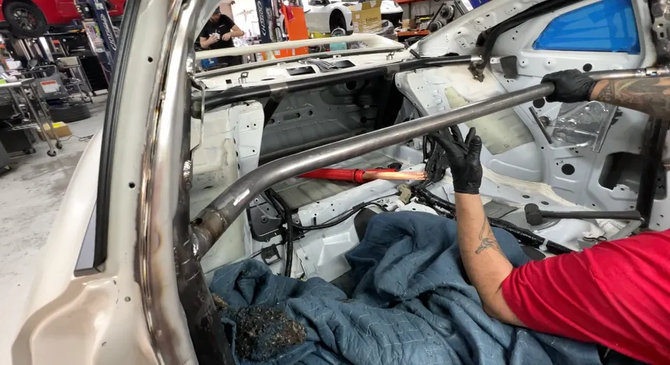 A mechanic installs a custom roll cage inside a white performance car at a tuning shop, with welding visible.