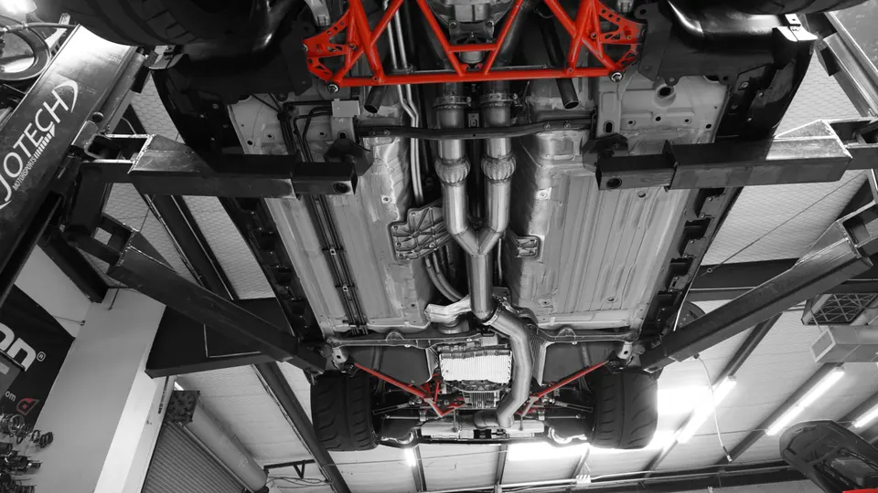 Underside view of a performance car on a lift, showcasing an aftermarket exhaust system and red chassis bracing in a tuning shop.