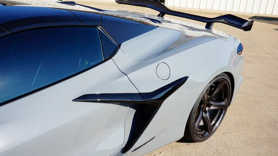 Close-up of a grey Chevrolet Corvette C8 rear quarter panel, showcasing a large black aftermarket rear wing, side air intake, and custom carbon fiber wheels.