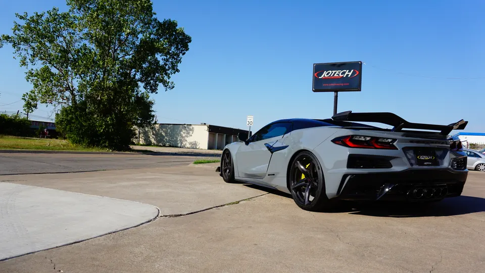 Grey Corvette C8.R with a large rear wing parked outside Jotech Performance shop on a sunny day.