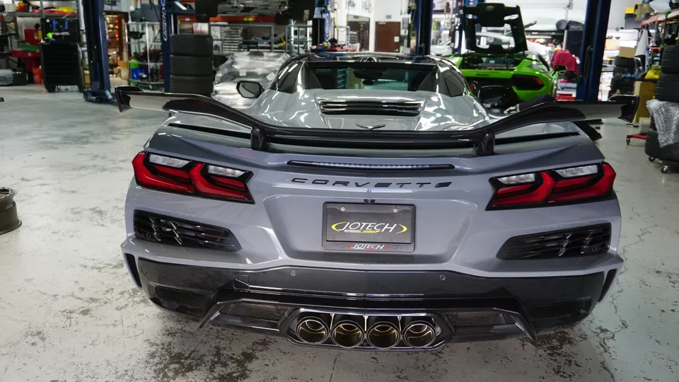 Rear view of a grey Chevrolet Corvette C8 with a large aftermarket rear wing and quad exhaust tips in a Jotech Motorsports shop.