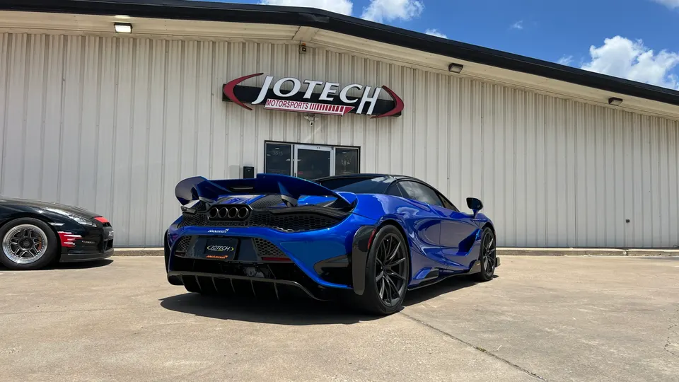 Blue McLaren 720S with an aftermarket wing and diffuser parked outside Jotech Motorsports, with a Nissan GT-R partially visible.