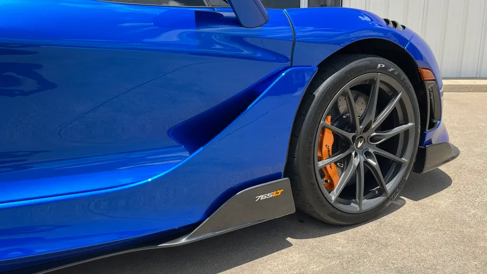 A close-up detail of a blue McLaren 765LT's front wheel, orange brake caliper, and carbon fiber side skirt.
