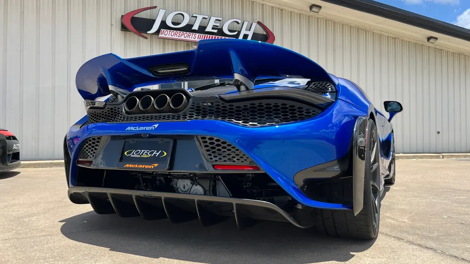 Rear view of a blue McLaren supercar with a large rear wing, quad exhaust, and aggressive diffuser parked outside Jotech Motorsports.