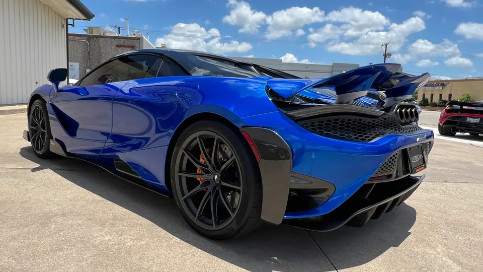 A vibrant blue McLaren supercar with an aggressive carbon fiber rear wing and diffuser, parked outdoors on a sunny day.