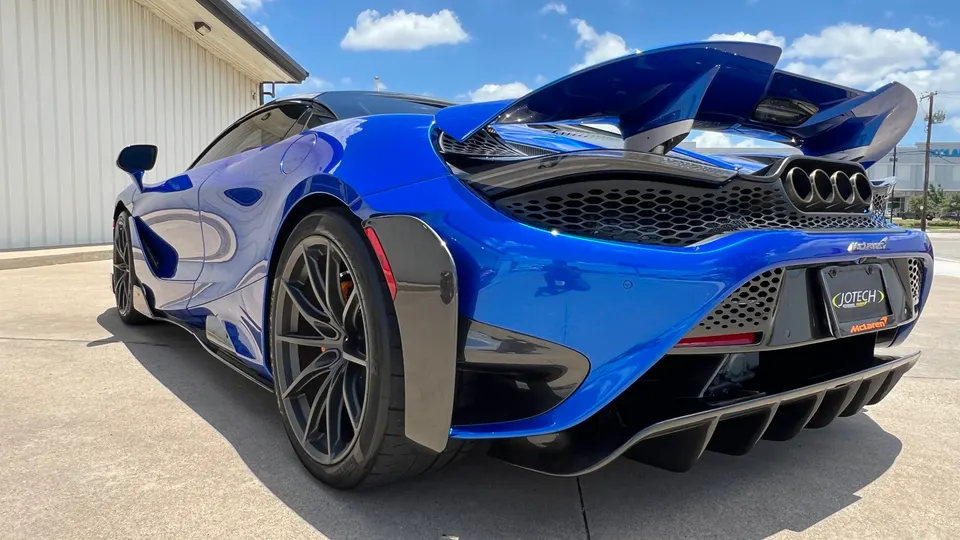 A vibrant blue McLaren supercar featuring a large rear wing, carbon fiber aero, and a prominent rear diffuser, parked outdoors with a Jotech license plate frame