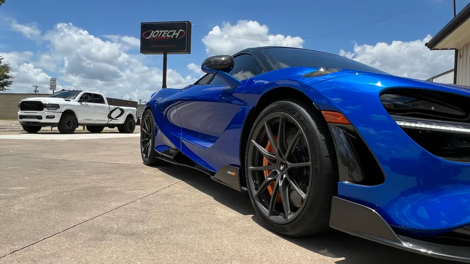 A vibrant blue McLaren 720S supercar parked outside Jotech Motorsports, with a white Ram truck in the background.