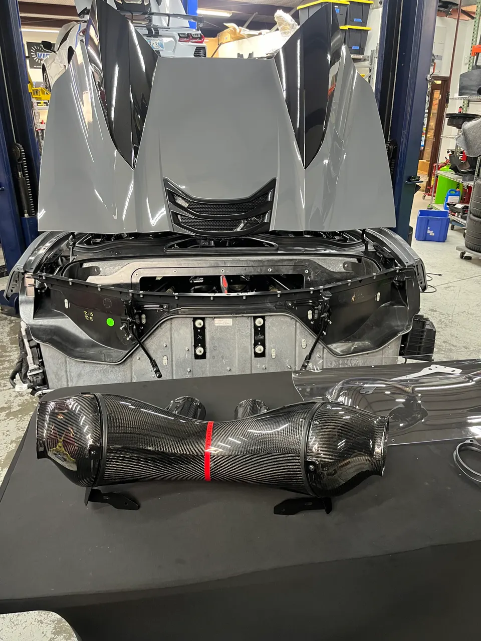 A grey Corvette C8 with its front hood open, revealing the engine bay, and a carbon fiber aftermarket intake system on a table in a performance shop.