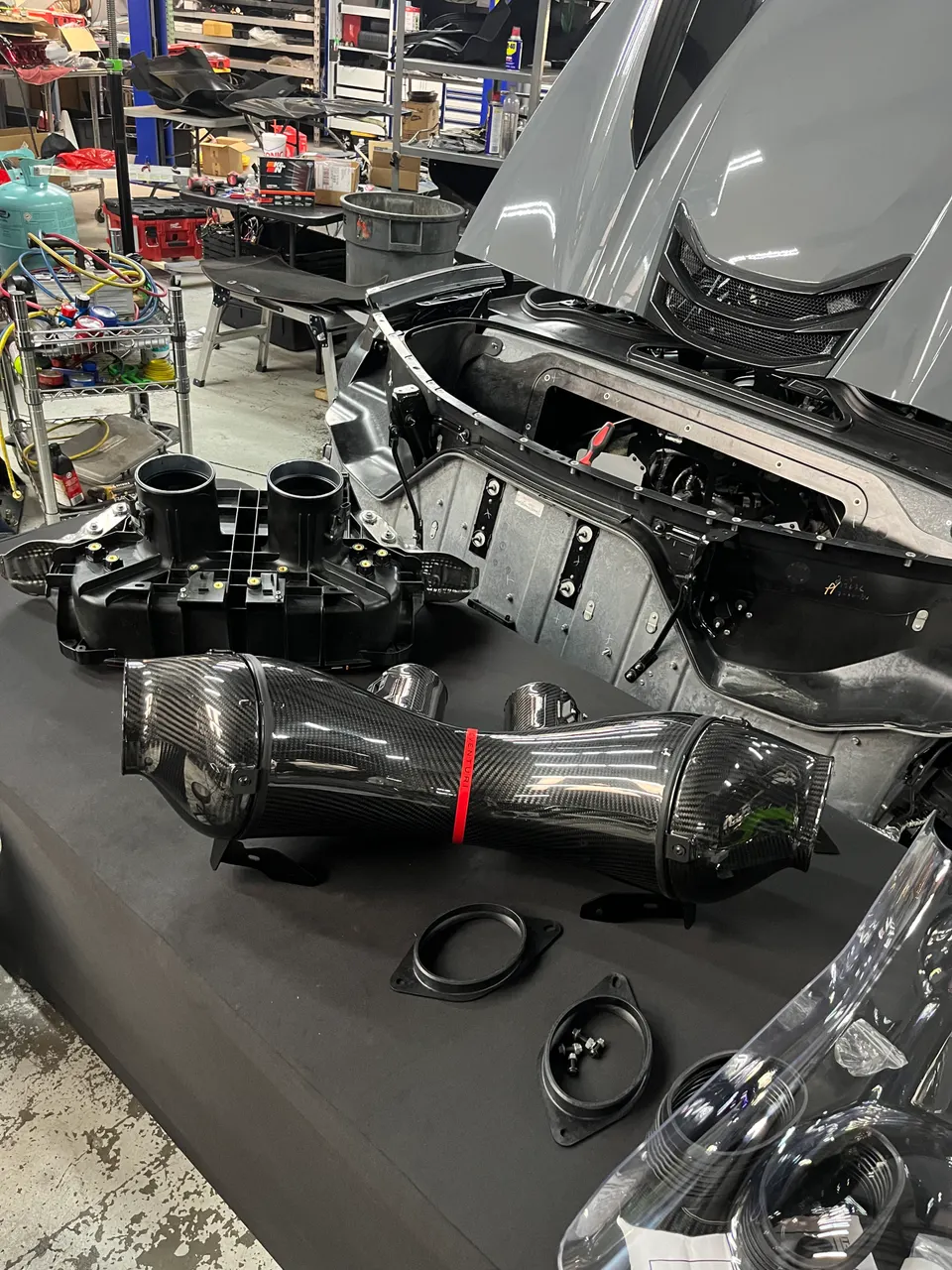 Carbon fiber intake system components laid out on a table in a performance shop, with a gray Corvette C8 in the background.