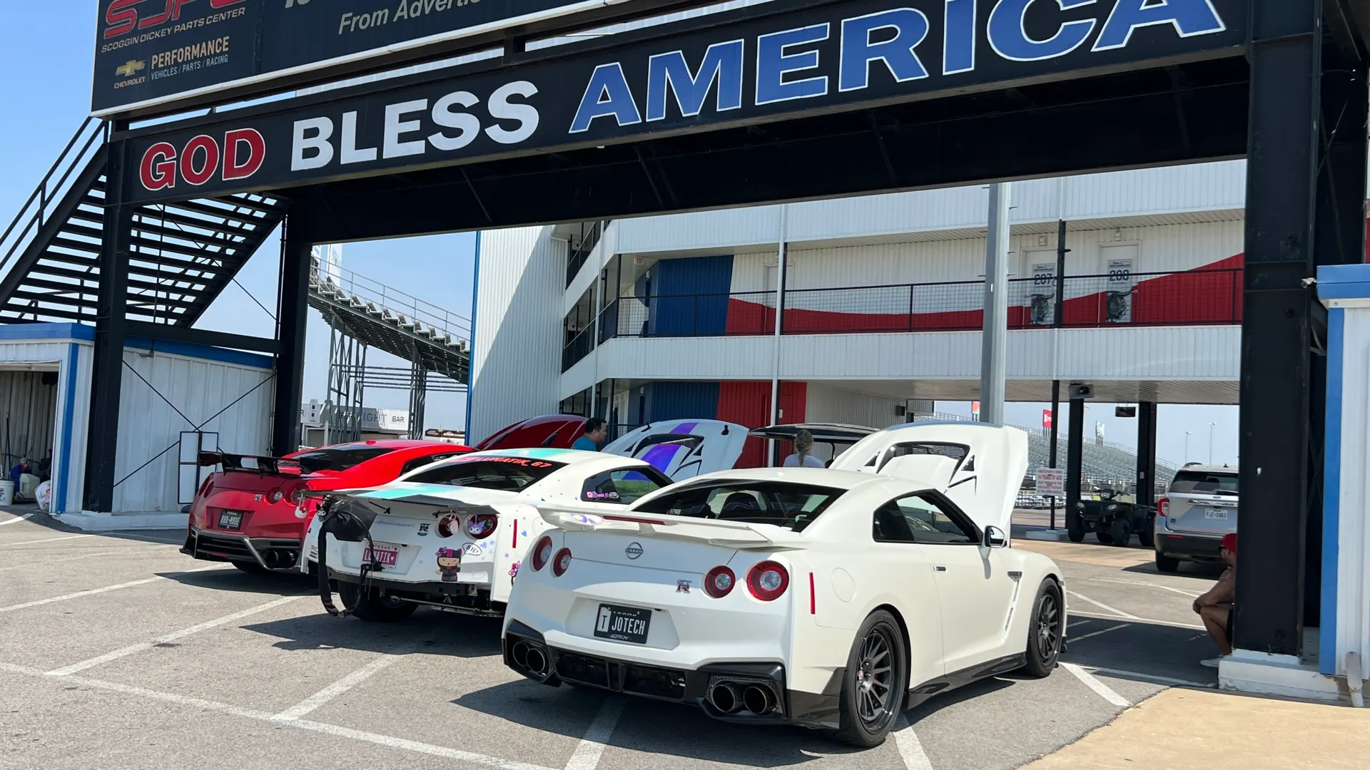 Several Nissan GT-R sports cars parked in a track paddock area under a "GOD BLESS AMERICA" banner.