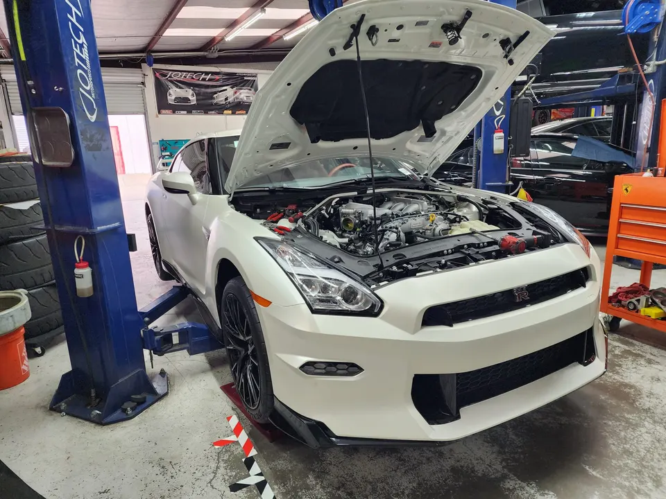 White Nissan GT-R on a blue car lift in a Jotech Motorsports shop with the hood open, revealing the engine bay.