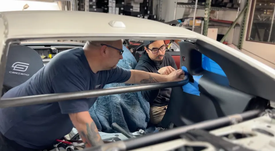 Two mechanics installing a roll cage in the interior of a car, with blue painter's tape visible during the process.