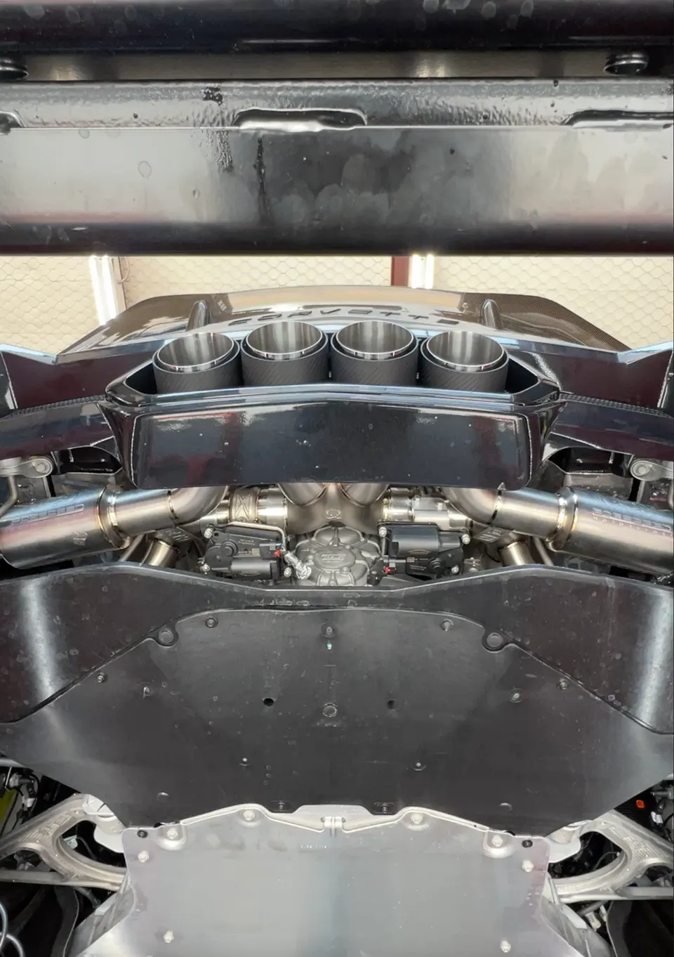 Close-up view of a Corvette's rear engine bay and performance exhaust system with quad tips, seen from below on a lift.