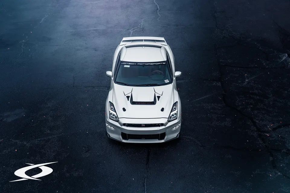 A white Nissan GT-R R35 with an aftermarket hood and aero, parked on dark asphalt, featuring a Jotech Motorsports banner.