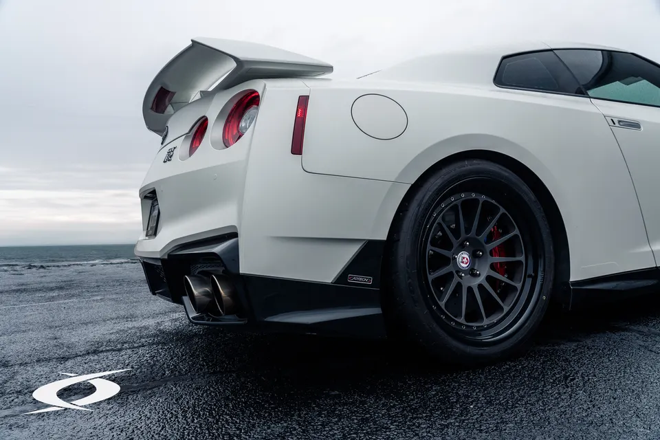 White Nissan GT-R with aftermarket HRE wheels, rear wing, and exhaust tips parked on a wet road with the ocean in the background.