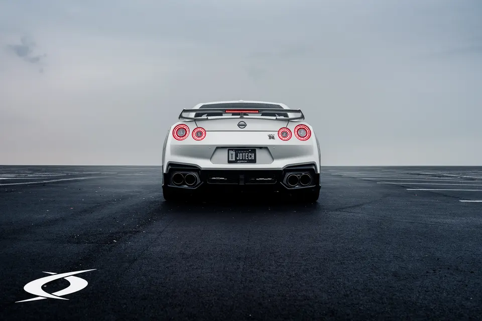 A white Nissan GT-R R35 from the rear, showcasing its quad exhaust tips and large rear wing on an open tarmac.