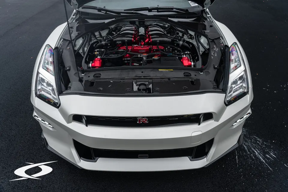 Front view of a white Nissan GT-R with its hood open, showcasing a highly modified engine bay with red intake manifolds.