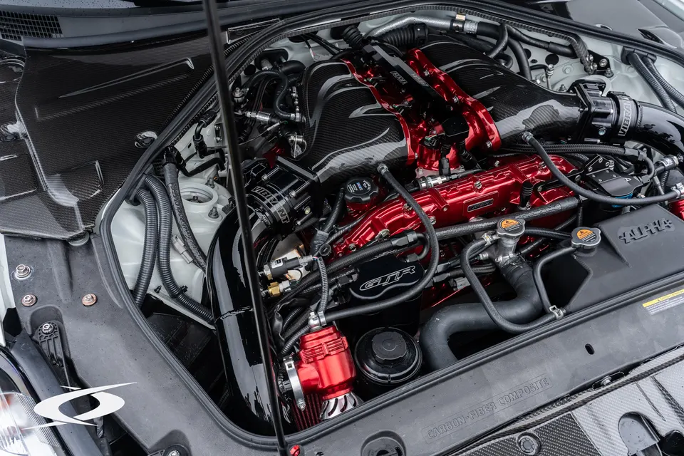 A detailed view of a custom Nissan GT-R engine bay featuring a red intake manifold, carbon fiber boost pipes, and a blow-off valve.